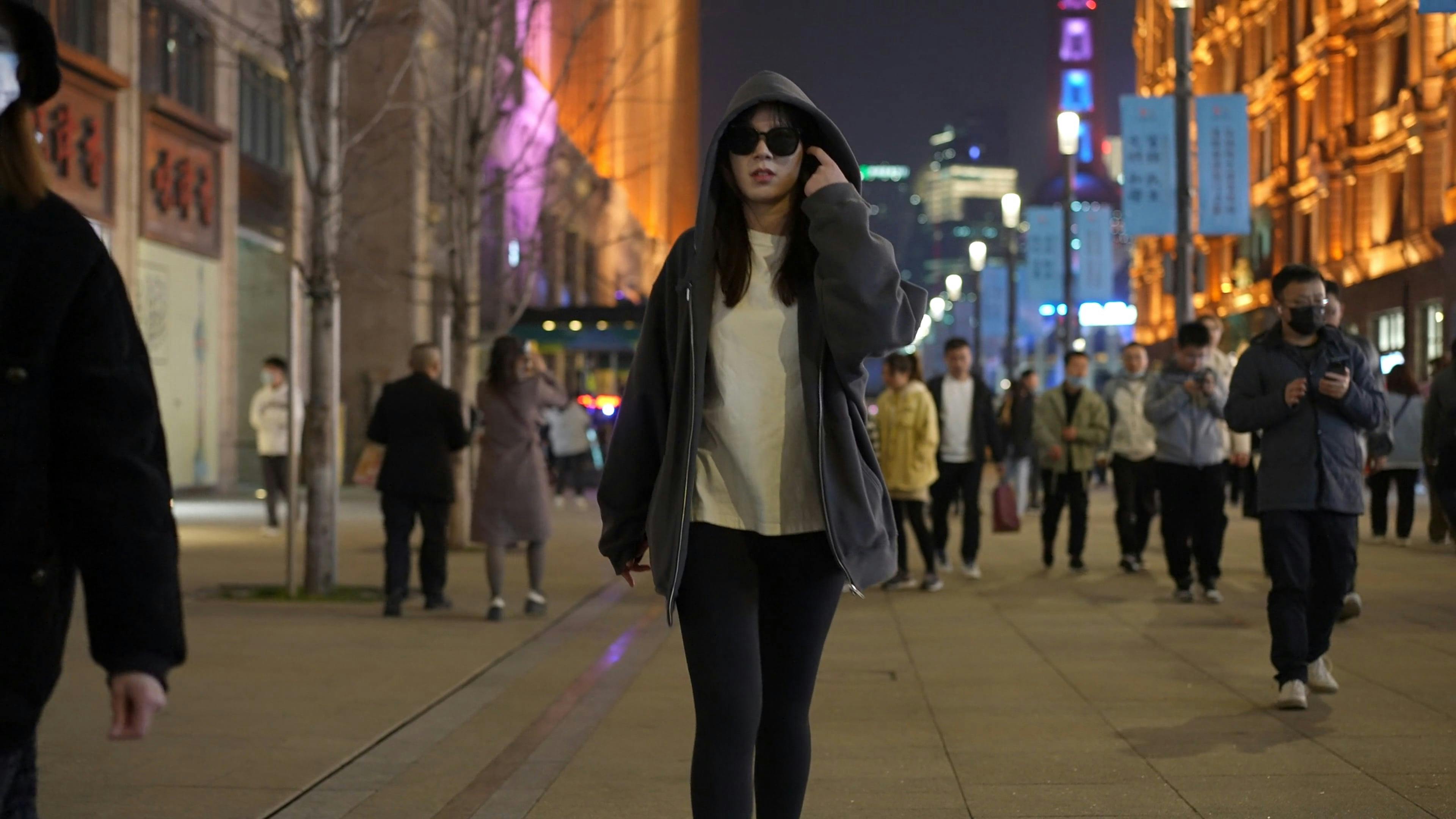 a woman in a hoodie and sunglasses walks down a city street 16379958