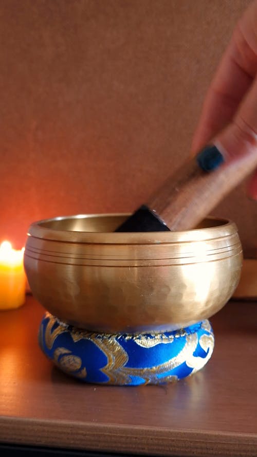 Close-up of woman hand using a Tibetan singing bowl placed on a ring cushion and burning incense and candle in the background. Hand rotation with wooden striker emits a relaxing vibration ...