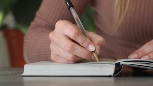 Close-up of woman sitting at a desk in the office and writing a note with a pen in a notebook. Selective focus technique on woman's hand