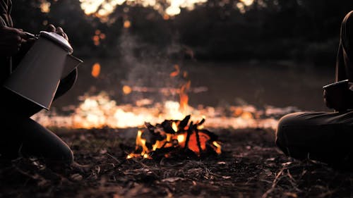 Two People In Front Of A Campfire Pouring Water From A Kettle