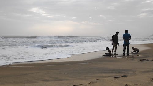 Dzieci Bawiące Się Na Piaszczystej Plaży W Pobliżu Dwóch Dorosłych Mężczyzn W Rozmowie, Stojąc