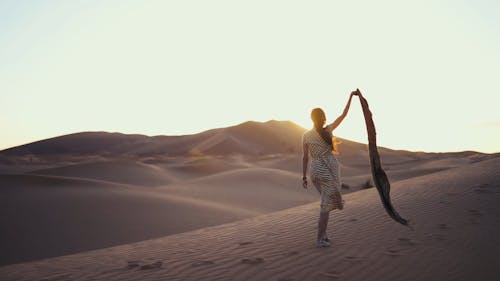 一個女人在大風天赤腳走在沙漠的沙子上舉起她的圍巾的手臂的慢動作鏡頭