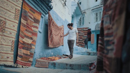 Slow Motion Footage Of A Man Walking Down The Steps Of An Alley Looking At Carpets Of Assorted Designs Hanging On Walls