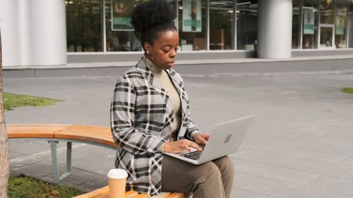 A Woman Seated On An Outdoor Bench Have A Sip Of Coffee While Working On Her Laptop