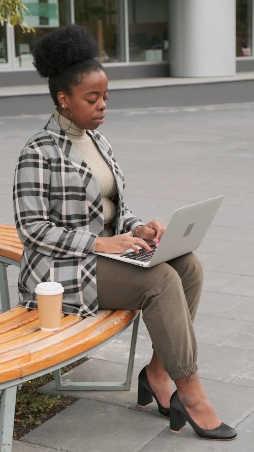 A Woman Seated On A Bench Drinks Her Coffee While Working On Her laptop