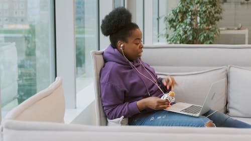 A Woman On A Video Call With Her Laptop Seated In A Sofa While Eating A Candy Bar