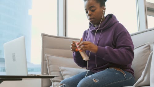 A Woman Seated In A Couch Working On Her Laptop Opens A Candy Bar To Eat