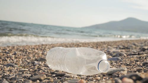 Close-up Of A Discarded Plastic Bottle On The Rocky Shore With The Waves Breaking In Slow Motion