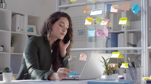 A Woman On Her Cellphone Inside An Office Looking And Sticking A Memo Stick Pad On A Glass Panel Besides Her Table