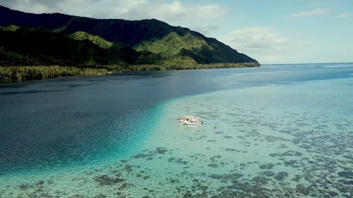 Aerial Footage Of People Swimming Around A Boat Docked In Shallow Crystal Clear Waters Near Beautiful Green Mountains