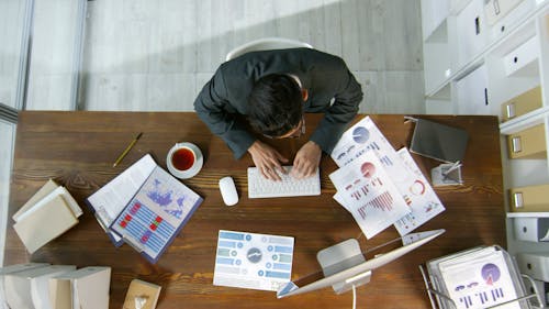 A Man Using His Computer To Record The Data On The Documents On His Desk