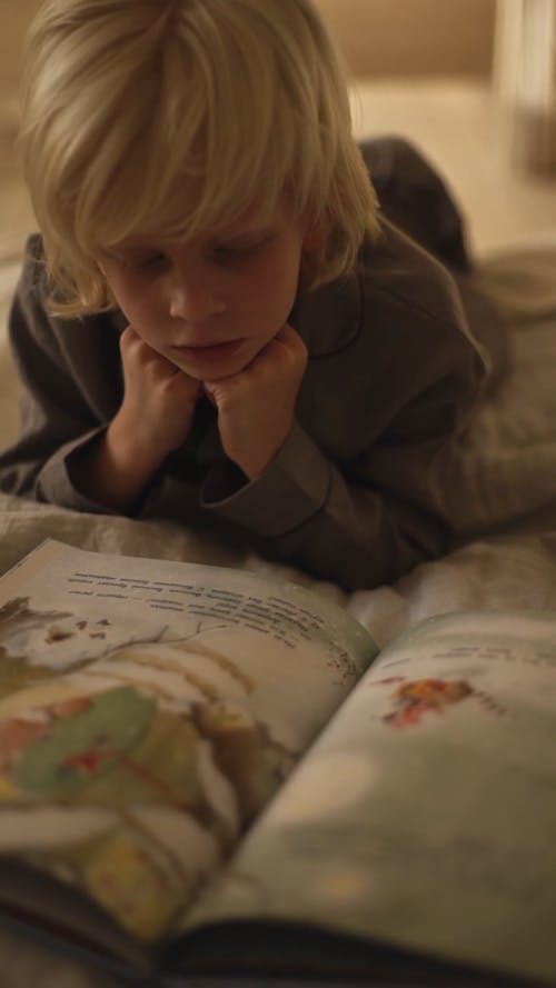 A Boy Reading A Book On A Bed