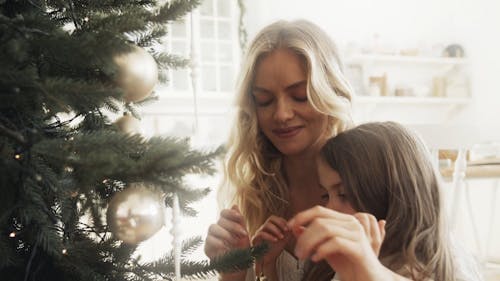 A Mother Assisting Her Daughter In Placing A Christmas Ball On A Christmas Tree