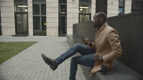 Man Sitting Outside A Building Looking At His Phone While Waiting