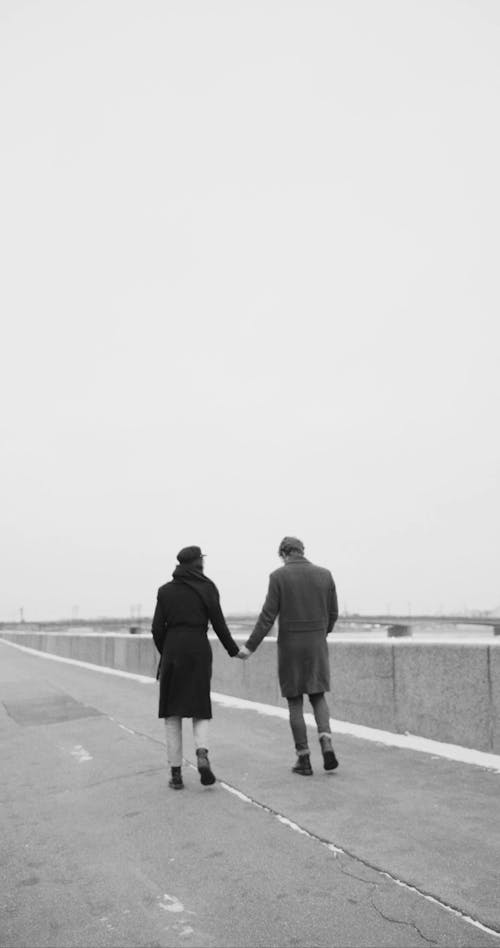 A Couple Walking Hand In Hand Along The Sidewalk By The Riverbank