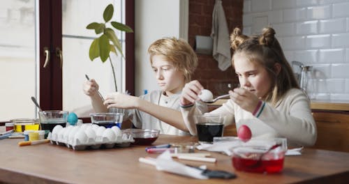 A Boy And A Girl Soaking White Eggs On Colored Liquids Using A Spoon