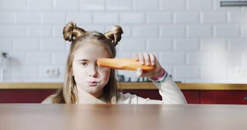 A Girl Dancing Behind The Kitchen Counter While Eating Fresh Peeled Carrots