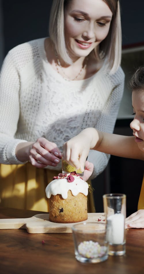 A Young Girl Sprinkle Topping On A Cake Guided By Her Mother