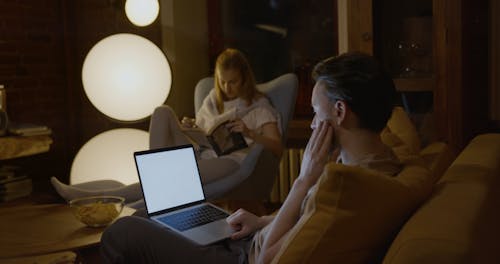 A Man Working On His Laptop While His Partner Is Reading A Book