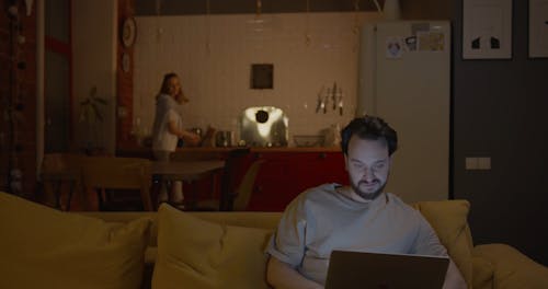 A Man Working On His Laptop With Her Partner Bringing Over Chips To Eat