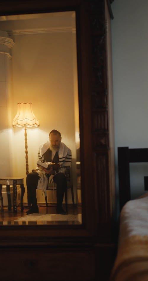 A Man Fixing His Tallit Using A Mirror After Reading A Book