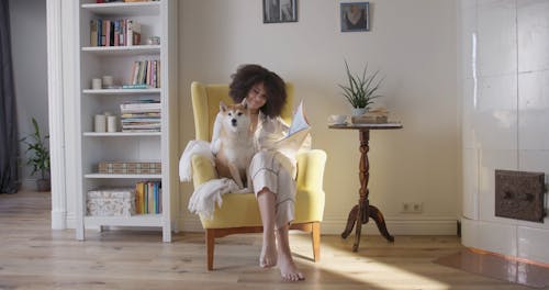 A Woman Having Coffee And Reading Magazine While Seated On A Chair With Her Dog