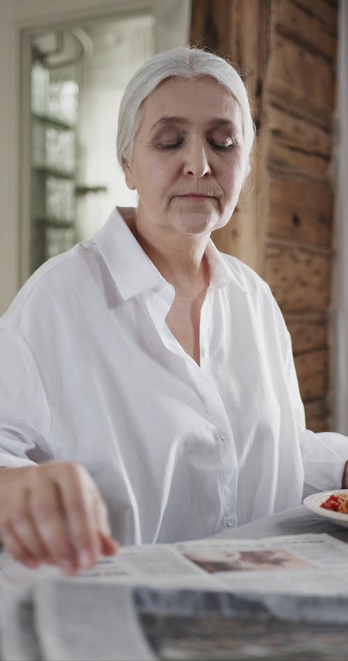 An Elderly Woman Reading A Newspaper In The Dining Table