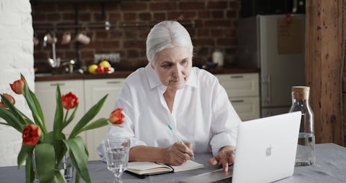 An Elderly Woman Using A Laptop And Taking Notes