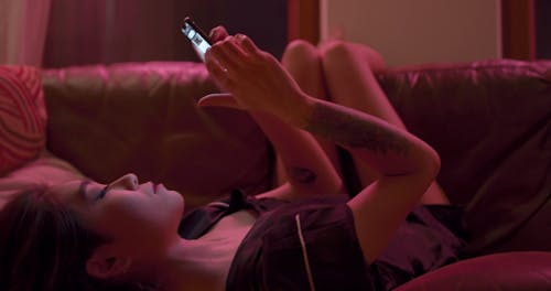 A Woman Lying Lazily On A Couch Looking Photos On Her Cellphone