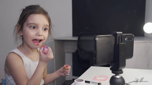 Little Girl Applying Lipstick While Looking At Her Smartphone