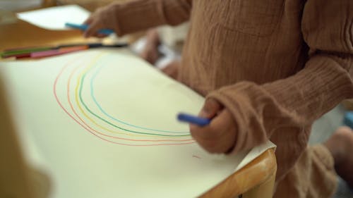 A Kid Drawing A Rainbow With Crayons On A Paper