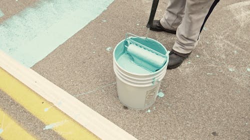 A Person Dipping A Paint Roller In A Bucket 