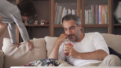 Dad Drinking From Plastic Cup While His Daughter Jumps on a Sofa