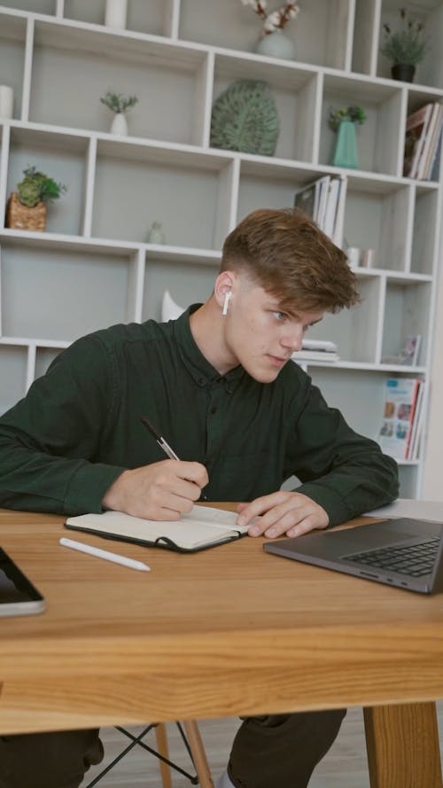 Man in Dark Green Long Sleeves Writing While Speaking Through the Laptop