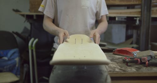 A Craftsman Shaping a Skateboard in Progress then Blowing Off the Wood Dust