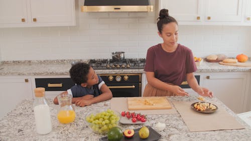 Woman Chopping Fruits in Kitchen and Son Standby 