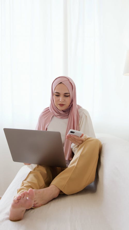 Woman in Pink Hijab Sitting on White Sofa While Using Her Laptop and Smartphone