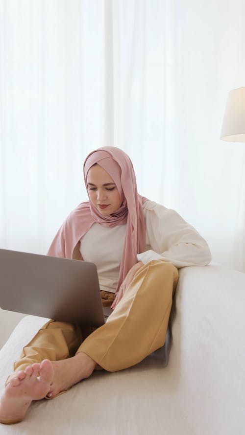 Woman in Pink Hijab Sitting on White Sofa While Using Her Laptop