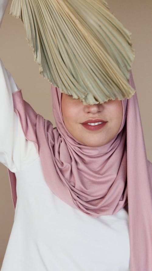 Woman in Pink Hijab Smiling While Covering Part of Her Face With a Leaf
