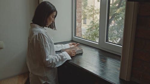 Woman in White Dress Shirt Holding a Bible and Cross While Praying by the Window