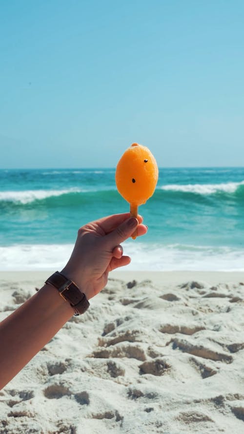 Person Holding an Orange Popsicle Against the Crashing Waves on the Beach