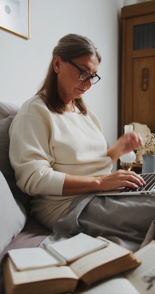 Woman Sitting on Her Bed While Using Her Laptop and Writing on a Notebook