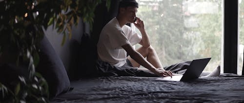 A Man Sitting on His Bed Using His Laptop While Having a Phone Call