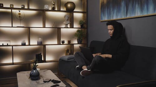 A Man in Black Hoodie Sitting on a Sofa While Busy Working on His Laptop