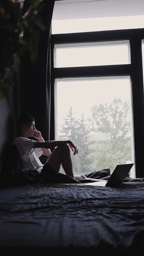 A Man Sitting on His Bed Using His Laptop While Having a Phone Call