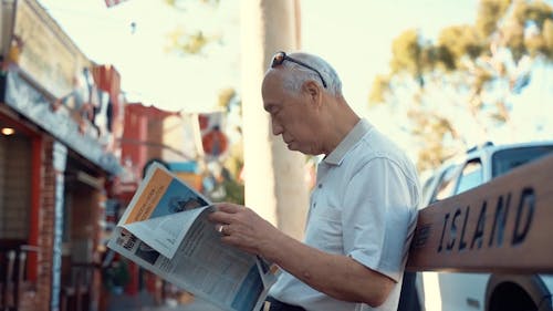 An Elderly Man Reading A Newspaper Seating In The Bench