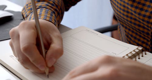 A Person Writing on His Notebook with a Pencil