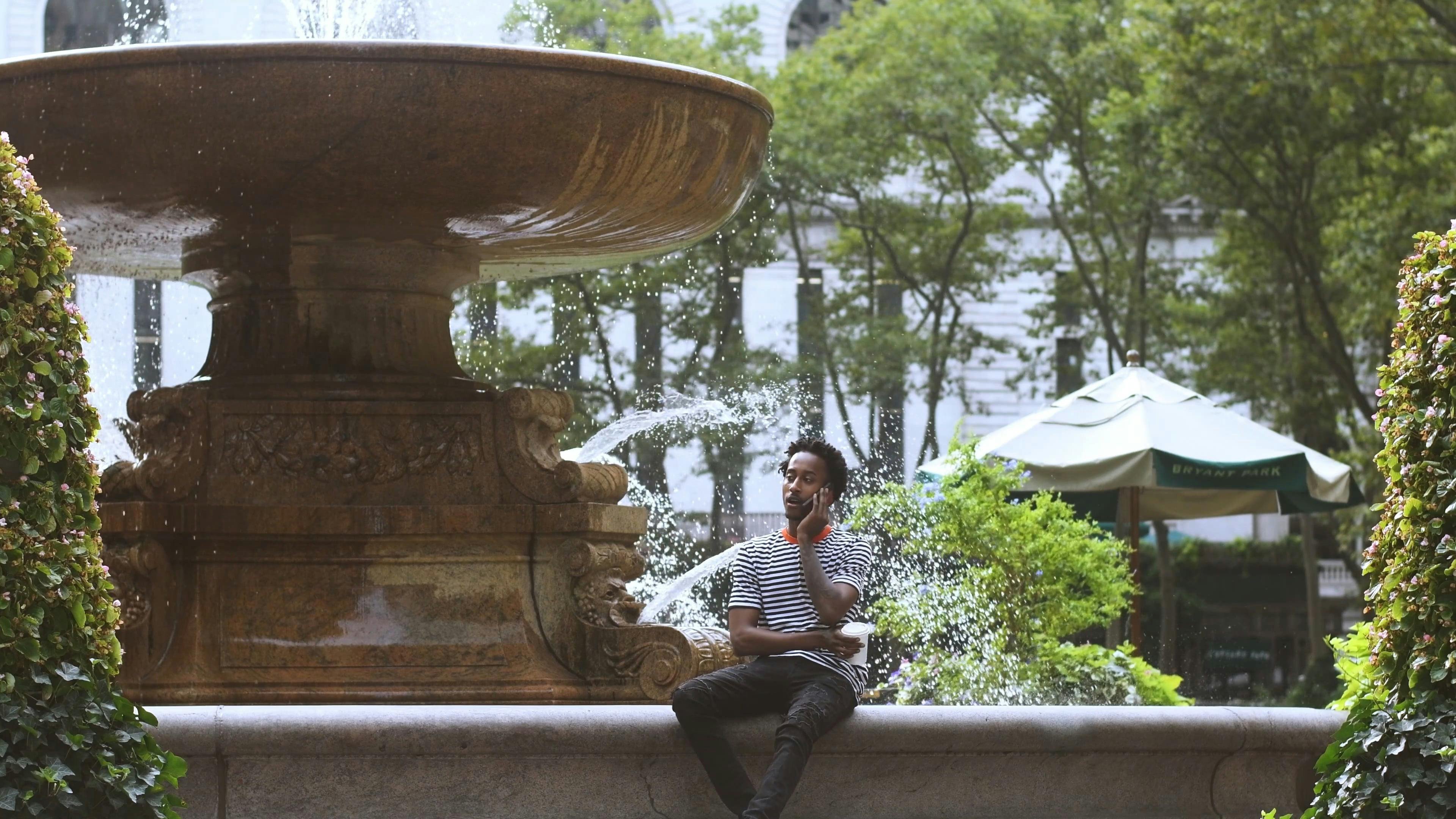 Man Talking on Phone While Sitting on a Water Fountain · Free Stock Video