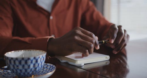 A Man Writing on a Notebook After Drinking on a Cup