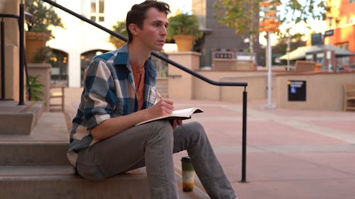 A Man Sitting on Concrete Stairs Writing in a Notebook